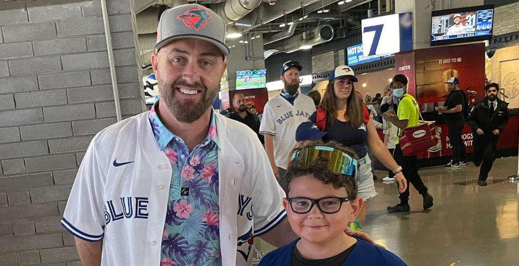 Realtor Kevin Kingma in a white BlueJays jersey smiles with his arms around a young boy with glasses in a BlueJays ball cap