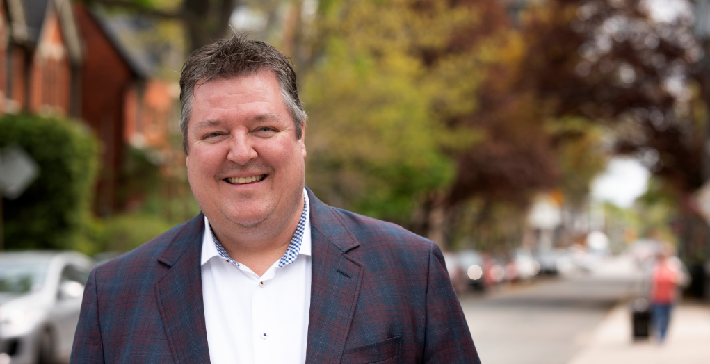 Chris stands smiling in a plaid blazer on a Toronto city street with lush treeswith tree