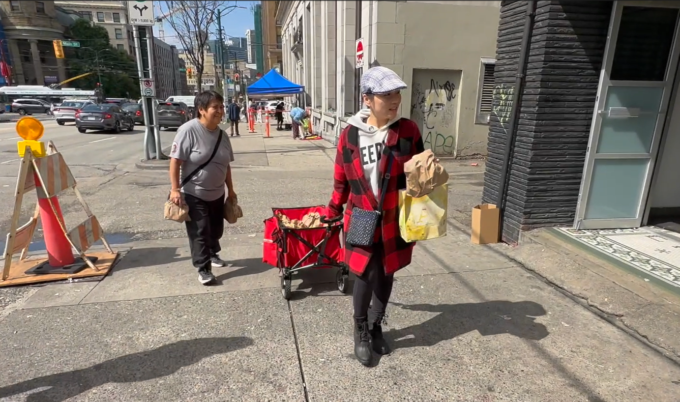 Crystal walking on a Downtown Eastside street pulling a red cart for the lunch program. In the background you can see unhoused people and a care tent.
