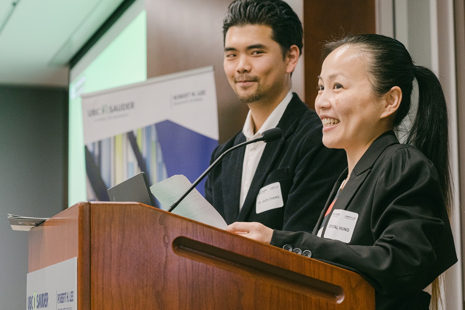 Crystal and her husband stand at a podium talking to a crowd after committing a 1-million-dollar donation. Behind them is a banner that says UBC Sauder.