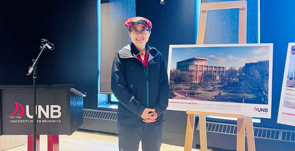 Li stands next to a University of New Brunswick Podium smiling. To her left is an artist mock-up of a new health centre.