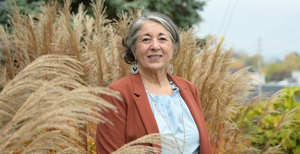 • Grace, le regard confiant, portant des boucles d’oreilles perlées d’inspiration autochtone et un veston, se tient debout parmi l’herbage naturel à l’extérieur de son agence.