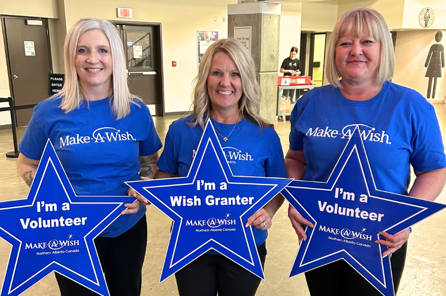 Lori is smiling and holding a sign that says 'I'm a Wish Granter'. On either side of her are volunteers also smiling. All of them are wearing Make-A-Wish shirts.