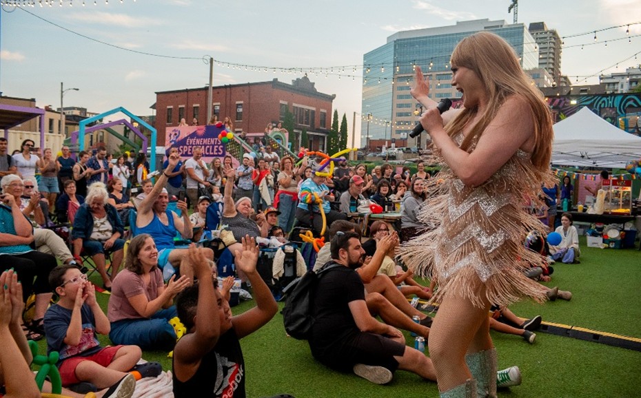 Community members sitting on grass, watching a drag queen singing