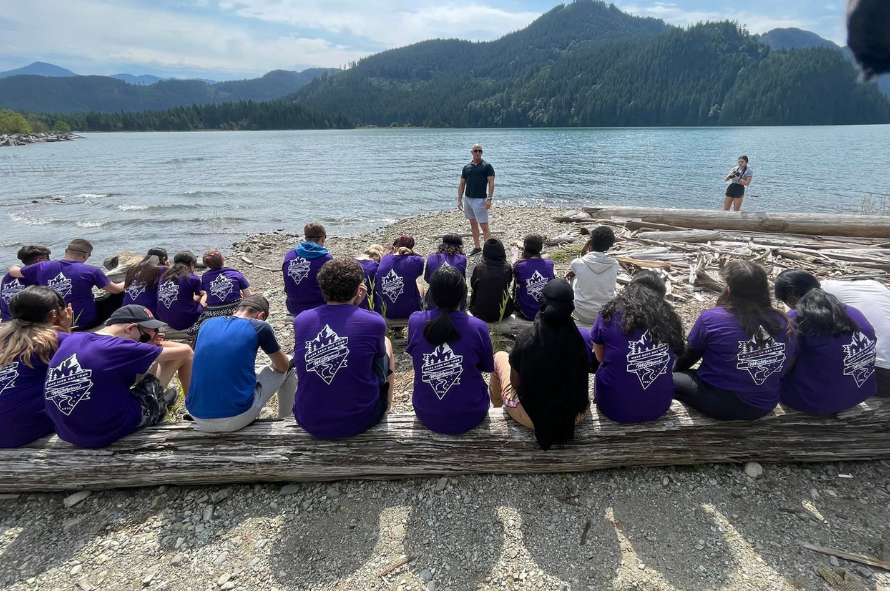 Teens sit on a beach listening to a John as he speaks -- Des adolescents sont assis sur une plage et écoutent John parler.
